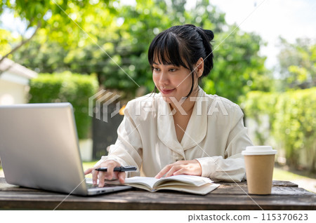 A positive Asian businesswoman working on her laptop computer at a wooden table in a garden. A positive Asian businesswoman working on her laptop computer at a wooden table in a garden. 115370623