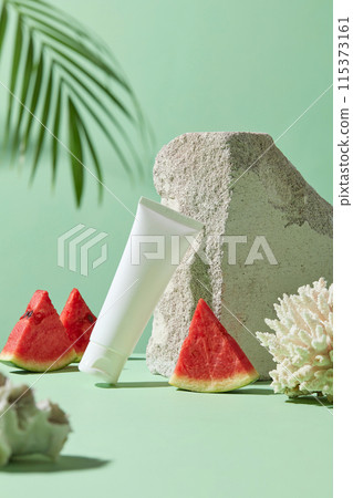 Cosmetic advertising photo on green background has watermelon ingredient. An empty label white tube leaning a large rock, decorated with white coral and green coconut leaf around, space for design Cosmetic advertising photo on green background has watermelon ingredient. An empty label white tube leaning a large rock, decorated with white coral and green coconut leaf around, space for design 115373161