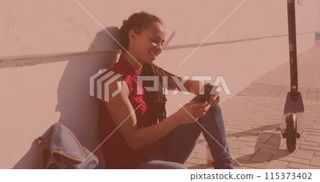 Biracial woman using a smartphone sitting on the side of the street 115373402