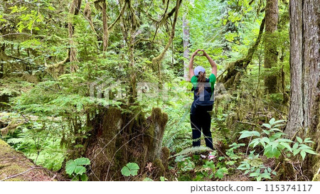 a girl in a Green T-shirt stands with her back in the jungle raised her hands up she shows her fingers the heart of a teenagers travel in Canada on Vancouver Island in a forest covered with moss ferns a girl in a Green T-shirt stands with her back in the jungle raised her hands up she shows her fingers the heart of a teenagers travel in Canada on Vancouver Island in a forest covered with moss ferns 115374117