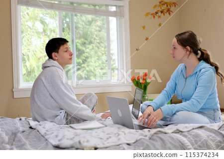 boy and girl teenagers sitting opposite each other with laptops working relaxing watching movies war teenagers gaze into the eyes against the background of the window in the room on the bed 115374234