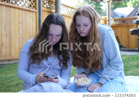 Two girls friends on a picnic drinking orange juice taking a selfie talking sitting on their phones sitting on the grass in summer dresses Two girls friends on a picnic drinking orange juice taking a selfie talking sitting on their phones sitting on the grass in summer dresses 115374263