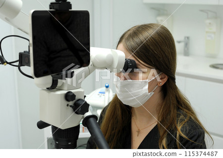 Big screen tablet young dentist sits near microscope Calipso scanner research prosthetics filling canals teeth long hair black dress code white background latest technology equipment microscope 115374487