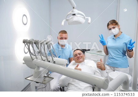 masks and gloves in blue color, transparent glasses in white clean stomatology Three joyful and contented patient, doctor, dental assistant show class dental treatment remove tartar in dental office masks and gloves in blue color, transparent glasses in white clean stomatology Three joyful and contented patient, doctor, dental assistant show class dental treatment remove tartar in dental office 115374533