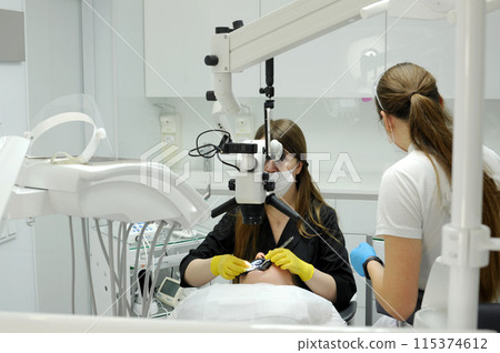A young girl on a dental chair and a dentist who sits next to him. He looks at the teeth with a dental microscope and holds a dental bur and a mirror in his hands. Dentist and patient during procedure 115374612