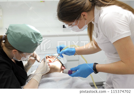 dentistry doctor treats a tooth in a man patient women in masks on them sterile gloves smart watch modern technology dental treatment tooth extraction hurt teeth cleaning teeth dental office 115374702