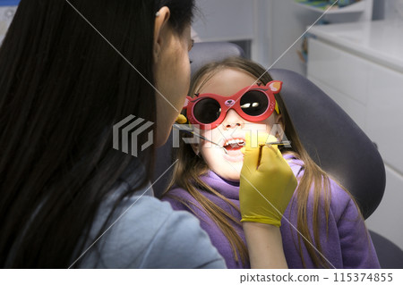 pediatric dentistry close-up little girl opened mouth shows teeth doctor looks at whether she has caries fillings treatment examination by doctor shine in eyes yellow hygienic gloves 115374855