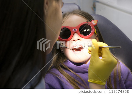 dental filling in children milk teeth deteriorate quickly dentist blows air on a caries hole and looks through it with a mirror Girl sits in a dental chair in sunglasses young beautiful real people dental filling in children milk teeth deteriorate quickly dentist blows air on a caries hole and looks through it with a mirror Girl sits in a dental chair in sunglasses young beautiful real people 115374868