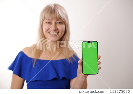 supply demand An adult woman 45-50 with blond hair in a blue dress is standing with mobile phone Green screen chroma key smiles advertising any products Sincere smile beautiful face blurred background supply demand An adult woman 45-50 with blond hair in a blue dress is standing with mobile phone Green screen chroma key smiles advertising any products Sincere smile beautiful face blurred background 115375030