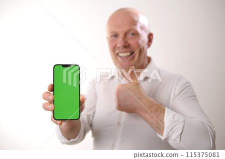 Elderly white man 60-65 years old happy and smile shows off something on smartphone with green screen to camera. Focusing on smartphone with chromakey Bald man in a white shirt 115375081