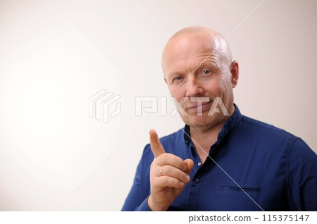 Emotions of an adult handsome bald man A man on a white background in a blue shirt The man smiles slyly and looks into the frame. He shows a thumbs up as if for some reason he wants to draw attention. Emotions of an adult handsome bald man A man on a white background in a blue shirt The man smiles slyly and looks into the frame. He shows a thumbs up as if for some reason he wants to draw attention. 115375147