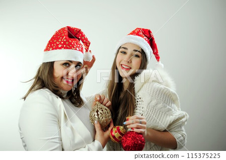 Mom and daughter are dressed in white with red New Year's hats on their heads holding Christmas toys many emotions of joy on a white background party invitation home celebration Mom and daughter are dressed in white with red New Year's hats on their heads holding Christmas toys many emotions of joy on a white background party invitation home celebration 115375225