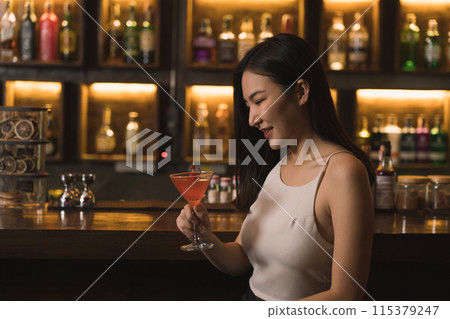 Asian woman drinking a cocktail at a bar at night. Asian woman drinking a cocktail at a bar at night. 115379247
