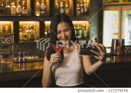 Asian woman taking a photo of herself while drinking whiskey at the bar. Asian woman taking a photo of herself while drinking whiskey at the bar. 115379248