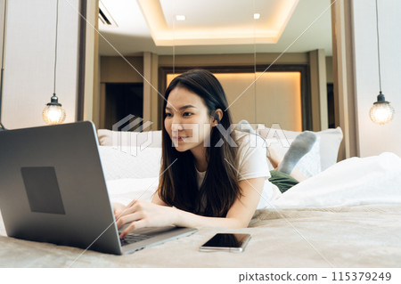 Asian woman laying on her laptop while enjoying herself in bed in her bedroom at home. Asian woman laying on her laptop while enjoying herself in bed in her bedroom at home. 115379249