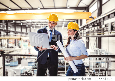 Two project managers standing in modern industrial factory. Manufacturing facility with robotics, robotic arms and automation. Storing products and materials in warehouse. 115381498