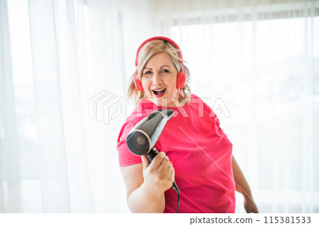 Portrait of beautiful young overweight woman at home, listening music via headphones, having fun, singing into hair dryer. 115381533