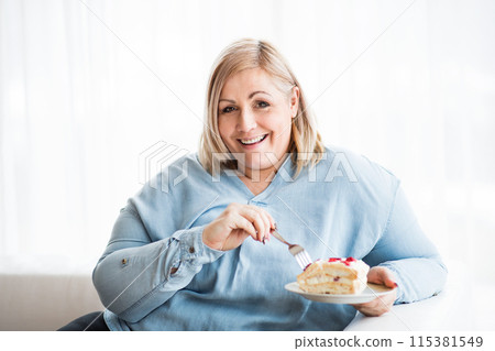 Portrait of beautiful young overweight woman at home, eating slice of cake, enjoying sweet taste. 115381549
