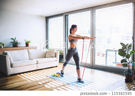 Beautiful redhead woman's home fitness routine with resistance band. Home workout on gym mt in living room, sporty woman wearing sportswear. 115381595
