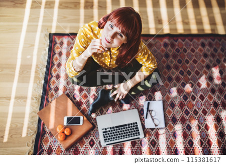 Redhead woman sitting on floor with laptop computer while working from home office. Businesswoman working remotely from her apartment in the city. High angle view, looking at camera. Redhead woman sitting on floor with laptop computer while working from home office. Businesswoman working remotely from her apartment in the city. High angle view, looking at camera. 115381617