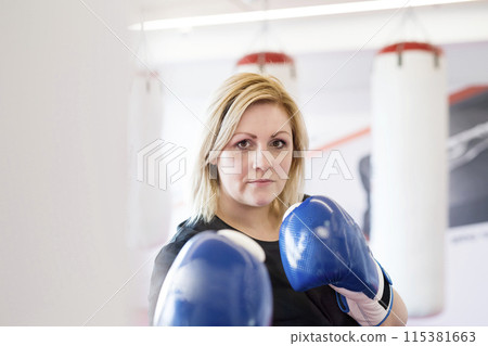 Overweight woman is working out in gym, boxing as part of her cardio training, wearing boxing gloves. Overweight woman is working out in gym, boxing as part of her cardio training, wearing boxing gloves. 115381663