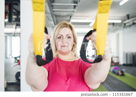 Overweight woman exercising in gym, doing suspension training, total resistance exercise. Personal trainer couching her and helping her. Overweight woman exercising in gym, doing suspension training, total resistance exercise. Personal trainer couching her and helping her. 115381667