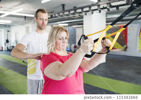 Overweight woman exercising in gym, doing suspension training, total resistance exercise. Personal trainer couching her and helping her. 115381668