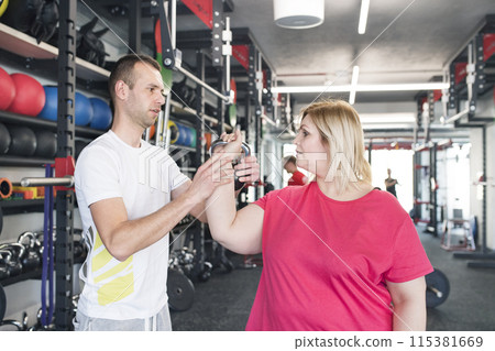 Overweight woman exercising in gym, using kettlebell. Personal trainer couching her and helping her. 115381669