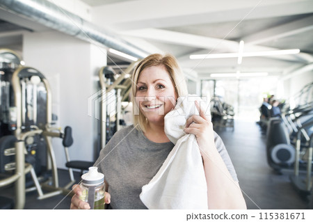 Overweight woman resting after workout in gym. Wipe sweat from face with towel. Overweight woman resting after workout in gym. Wipe sweat from face with towel. 115381671