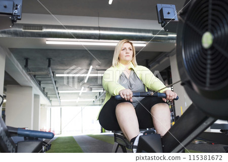 Overweight woman exercising on rowing machine. Routine workout for physical and mental health 115381672