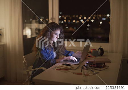 Beautiful university student is writing her thesis on a laptop at night, sitting at home at table, focusing on her research. Preparing for final exam, studying. 115381691