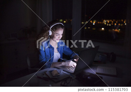 Beautiful university student is writing her thesis on a laptop at night, sitting at home at table, focusing on her research. Preparing for final exam, studying. Beautiful university student is writing her thesis on a laptop at night, sitting at home at table, focusing on her research. Preparing for final exam, studying. 115381703
