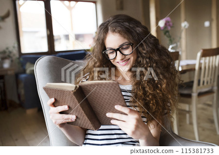 Female student sitting in armchair, reading book, studying for final exam at university. Beautiful woman relaxing at home. Female student sitting in armchair, reading book, studying for final exam at university. Beautiful woman relaxing at home. 115381733