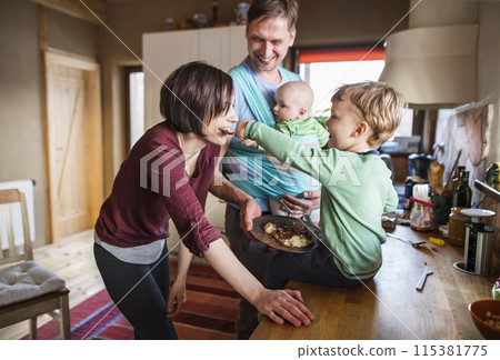Father holding small baby in baby sling. Older brother eating porridge on kitchen counter, feeding mother. Unconditional parental love, family moment. 115381775