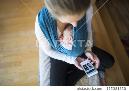 Newborn baby sleeping in baby sling, wrap, mother scrolling on smartphone. Unconditional paternal love, mother's Day. 115381850