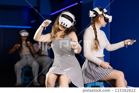 Girls wearing virtual reality glasses play in pairs, against each other, manipulating objects or shooting at targets using joysticks in their hands 115382622