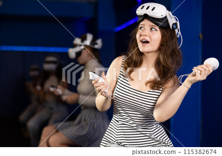 Girl gamer with virtual reality glasses on her forehead expresses enthusiastic emotions from playing with her friends in dark room Girl gamer with virtual reality glasses on her forehead expresses enthusiastic emotions from playing with her friends in dark room 115382674