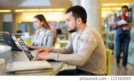 Focused young student studying with books and laptop in library 115382693