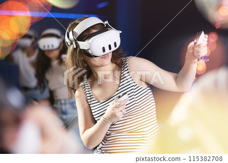 Portrait of young girlfriends using virtual reality goggles controlling joysticks sitting in room with friends Portrait of young girlfriends using virtual reality goggles controlling joysticks sitting in room with friends 115382708