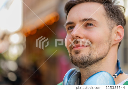 Thoughtful happy smiling male tourist with wireless headphones around neck looking up on city street Thoughtful happy smiling male tourist with wireless headphones around neck looking up on city street 115383544