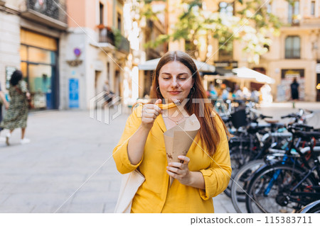 Carefree woman eating traditional Churros, a fried pastry with chocolate on a city street. Positive cheerful female student posing outdoors. Food concept Carefree woman eating traditional Churros, a fried pastry with chocolate on a city street. Positive cheerful female student posing outdoors. Food concept 115383711