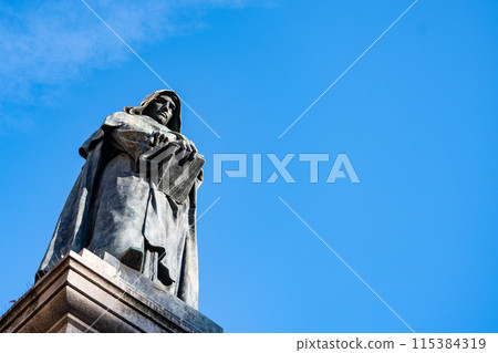 The Monument to Giordano Bruno, a bronze statue of the Italian philosopher holding a book, stands tall against a vibrant blue sky in Campo de Fiori, Rome. 115384319