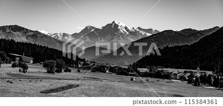 A panoramic summer vista of the Ortles Mountain surrounded by Alpine meadows, with a quaint village nestled in the foreground. Black and white photography. A panoramic summer vista of the Ortles Mountain surrounded by Alpine meadows, with a quaint village nestled in the foreground. Black and white photography. 115384361