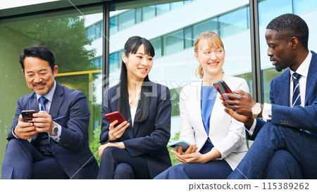 A group of multinational business people sitting on an outdoor bench and looking at their smartphones 115389262