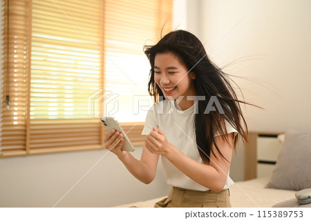 Young Asian woman reading text message on her phone with happy pleased face expression Young Asian woman reading text message on her phone with happy pleased face expression 115389753