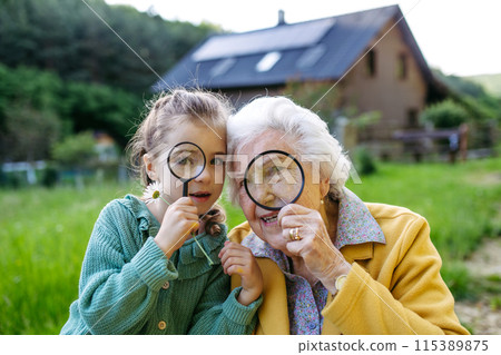 Granddaughter exploring wildlife with elderly grandma, using magnifying glass to look at insect. Senior lady spending time with young girl, enjoying together time. Granddaughter exploring wildlife with elderly grandma, using magnifying glass to look at insect. Senior lady spending time with young girl, enjoying together time. 115389875