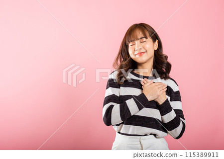 A happy woman in casual attire holds her hands close to her chest, meditating and smiling. Studio shot isolated on a pink background, portraying the concepts of belief, faith, and gratitude. 115389911