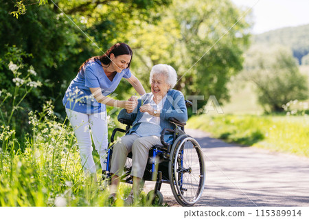 Female caregiver and senior woman in wheelchair picking wild flowers. Nurse and elderly woman enjoying a warm day in nursing home, public park. 115389914