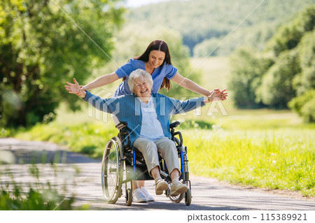 Female caregiver pushing senior woman in wheelchair. Nurse and elderly woman enjoying a warm day in nursing home, public park. Female caregiver pushing senior woman in wheelchair. Nurse and elderly woman enjoying a warm day in nursing home, public park. 115389921