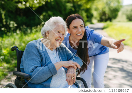 Female caregiver pushing senior woman in wheelchair. Nurse and elderly woman enjoying a warm day in nursing home, public park. 115389937
